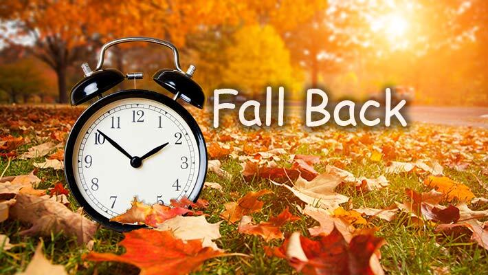 An alarm clock sitting in leaves with the text, Fall Back.